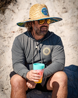 A man wearing a Blueline Signature straw lifeguard hat on the sand. Wide-brim sun protection with a chin strap for the beach in Jupiter, FL.