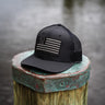Black trucker hat with a blacked-out American flag patch featuring a blue star field, sitting on a dock piling