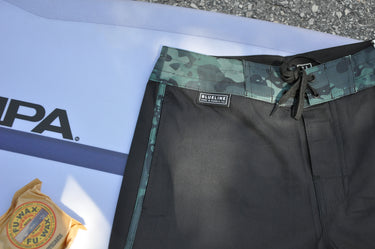 Black board shorts with green camouflage waistband on a white surface with 'Blueline' branding.