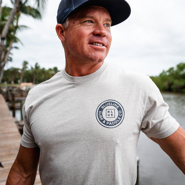 Man wearing a white t-shirt with a logo and a cap, standing by a body of water.