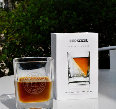 A clear whiskey wedge glass filled with amber liquid sits beside its white retail box on a white table outdoors, with green foliage in the background. The box is branded CORKCICLE.