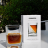 A clear whiskey wedge glass filled with amber liquid sits beside its white retail box on a white table outdoors, with green foliage in the background. The box is branded CORKCICLE.