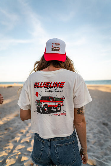 Blueline Women’s Christmas Crop T-Shirt 2025 Natural worn on Jupiter beach with holiday truck graphic – back view