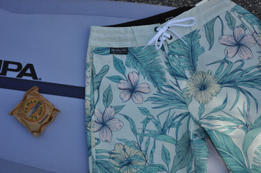 Floral-patterned board shorts next to a surfboard with Blueline Surf and Paddle label.