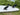 A white surfboard lying on grass outdoors, with three black surfboard fins attached. The fins have white printed logos and a small white recycling symbol.
