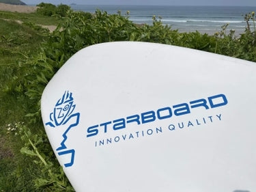 A white Starboard surfboard with blue branding, resting outdoors on green grass with the ocean and shoreline visible in the background.