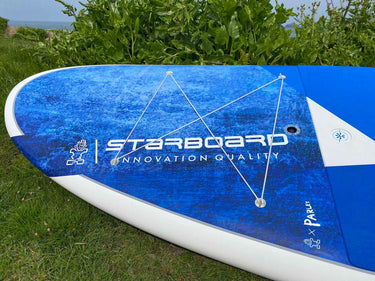A blue and white stand-up paddleboard photographed outdoors on grass with green foliage and water in the background. The board has visible branding including STARBOARD and Paralyo, with white rope leading attached across the deck.