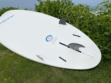A white surfboard with black fins and a blue logo, resting on green grass with leafy plants and water in the background.