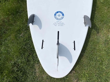 A white surfboard viewed from above, resting on green grass. The board features a blue circular logo in the center, black fin hardware, and two dark gray fins mounted on the sides.