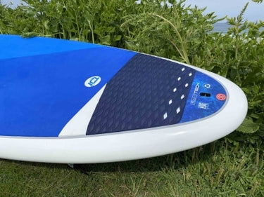 A blue and white stand-up paddleboard with a dark textured grip pad, photographed outdoors on grass with green foliage and water in the background.
