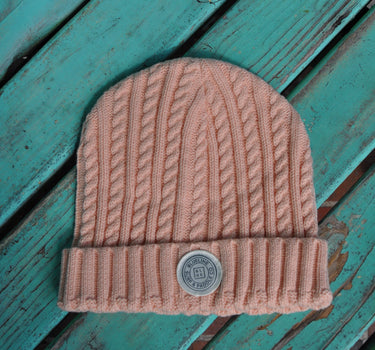 A dusty pink cable-knit baby beanie with a silver metal button closure, laid flat on a distressed teal wooden surface.