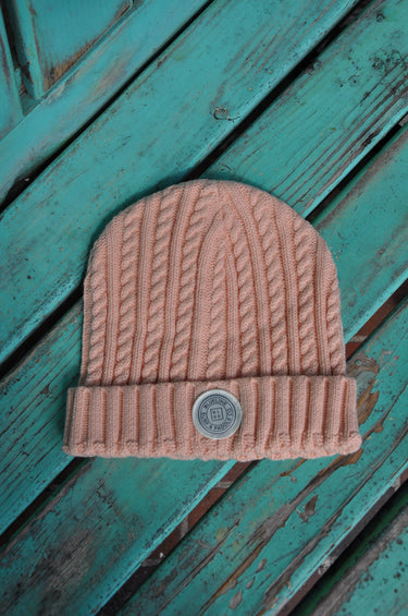 A dusty pink cable-knit baby beanie with a silver metal button closure, laid flat on a distressed teal wooden surface.