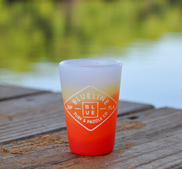A silicone shot glass with a Blueline Surf and Paddle diamond logo in marigold. Sitting on a dock in Palm Beach County. 