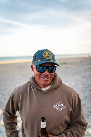 Man wearing a OG Blueline pale indigo navy coppertone hat and sunglasses, holding a beer bottle on a beach in a Blueline Surf and Paddle Hoodie