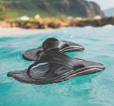 Olukai Ohana Black sandals floating in clear blue water.