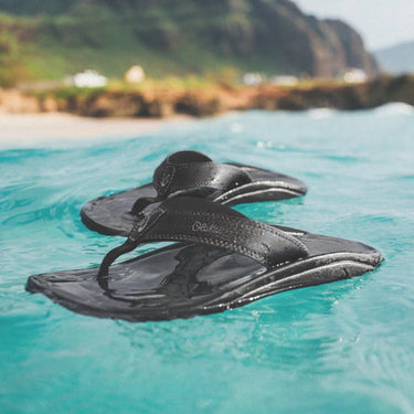 Olukai Ohana Black sandals floating in clear blue water.