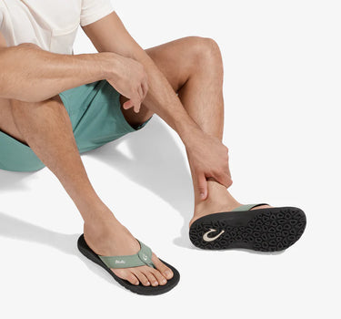 Man wearing OluKai ʻOhana seagreen balck sandals while relaxing, showcasing premium comfort and water-friendly performance.