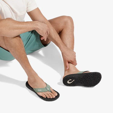 Man wearing OluKai ʻOhana seagreen balck sandals while relaxing, showcasing premium comfort and water-friendly performance.
