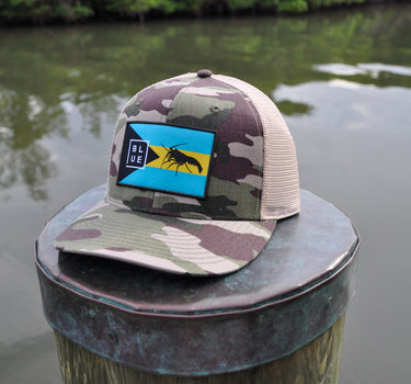 Camo Bahamas flag trucker hat by Blueline Surf & Paddle photographed on a weathered dock piling overlooking calm waterfront waters in Jupiter Florida.