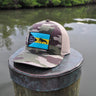 Camo Bahamas flag trucker hat by Blueline Surf & Paddle photographed on a weathered dock piling overlooking calm waterfront waters in Jupiter Florida.