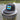 Black Bahamas flag trucker hat by Blueline Surf & Paddle photographed on a weathered dock piling overlooking calm Jupiter Florida waterfront waters.