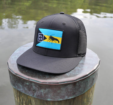 Black Bahamas flag trucker hat by Blueline Surf & Paddle photographed on a weathered dock piling overlooking calm Jupiter Florida waterfront waters.