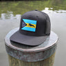 Black Bahamas flag trucker hat by Blueline Surf & Paddle photographed on a weathered dock piling overlooking calm Jupiter Florida waterfront waters.