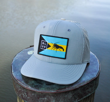 Gray Bahamas flag trucker hat by Blueline Surf & Paddle photographed on a dock piling overlooking calm waterfront waters in Jupiter Florida.