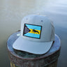 Gray Bahamas flag trucker hat by Blueline Surf & Paddle photographed on a dock piling overlooking calm waterfront waters in Jupiter Florida.