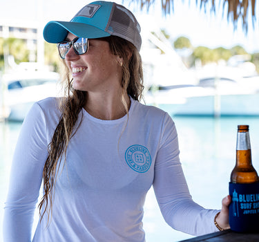 Woman wearing Blueline Surf & Paddle UV shirt and blue hat holding a Blueline Jupiter Lighthouse koozie on the Intracoastal Waterway in Jupiter, Florida, showcasing South Florida surf and paddle lifestyle apparel.