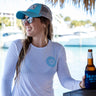Woman wearing Blueline Surf & Paddle UV shirt and blue hat holding a Blueline Jupiter Lighthouse koozie on the Intracoastal Waterway in Jupiter, Florida, showcasing South Florida surf and paddle lifestyle apparel.