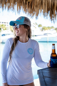 Woman wearing Blueline Surf & Paddle UV shirt and blue hat holding a Blueline Jupiter Lighthouse koozie on the Intracoastal Waterway in Jupiter, Florida, showcasing South Florida surf and paddle lifestyle apparel.