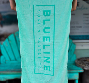 Blueline Beach Towel in mint green with vertical Blueline logo displayed outdoors at Blueline Surf & Paddle Co. in Jupiter, Florida.