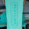 Blueline Beach Towel in mint green with vertical Blueline logo displayed outdoors at Blueline Surf & Paddle Co. in Jupiter, Florida.