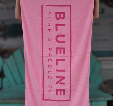 Blueline Beach Towel in pink featuring the vertical Blueline logo, displayed outside Blueline Surf & Paddle Co. in Jupiter, Florida.