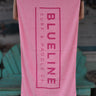 Blueline Beach Towel in pink featuring the vertical Blueline logo, displayed outside Blueline Surf & Paddle Co. in Jupiter, Florida.