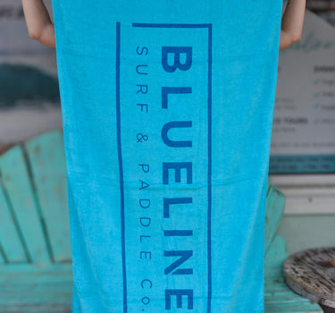 Blueline Beach Towel in turquoise featuring the vertical Blueline logo displayed outside Blueline Surf & Paddle Co. in Jupiter, Florida.