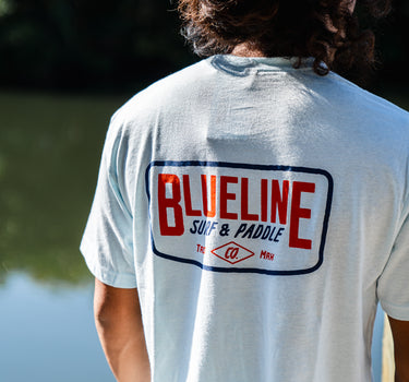 Back view of a man wearing an ice blue Blueline Classic Badge T-shirt with large back logo, standing near the water in Jupiter, Florida.