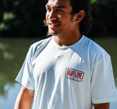 Man wearing an ice blue Blueline Classic Badge T-shirt with small chest logo, photographed outdoors near the water in Jupiter, Florida.