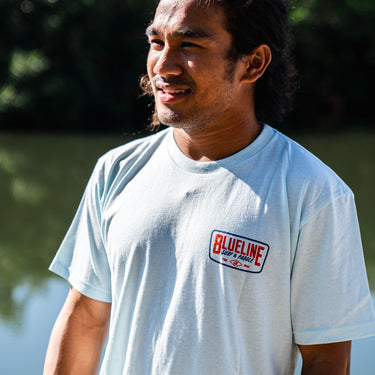 Man wearing an ice blue Blueline Classic Badge T-shirt with small chest logo, photographed outdoors near the water in Jupiter, Florida.