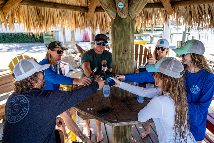 Group of friends wearing Blueline Surf & Paddle apparel sharing a toast under a tiki hut in Jupiter, Florida. Celebrating the South Florida lifestyle that inspires the Blueline brand.