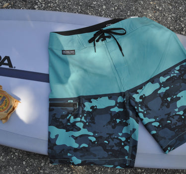Blueline Firefly performance boardshorts in Light Blue and Camo Slate laid across a surfboard.