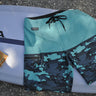 Blueline Firefly performance boardshorts in Light Blue and Camo Slate laid across a surfboard.