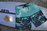 Blueline Firefly performance boardshorts in Light Blue and Camo Slate laid across a surfboard.