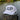 A white trucker-style cap with large dark gray 'JUP' lettering and 'BLUELINE SURF' text on the front, resting on a weathered wooden dock post with water and green trees in the background.