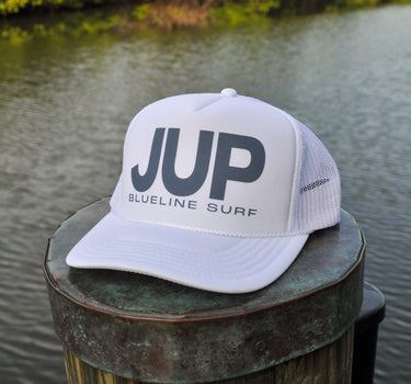 A white trucker-style cap with large dark gray 'JUP' lettering and 'BLUELINE SURF' text on the front, resting on a weathered wooden dock post with water and green trees in the background.