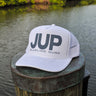 A white trucker-style cap with large dark gray 'JUP' lettering and 'BLUELINE SURF' text on the front, resting on a weathered wooden dock post with water and green trees in the background.