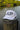 A white trucker-style cap with large dark gray 'JUP' lettering and 'BLUELINE SURF' text on the front, resting on a weathered wooden dock post with water and green trees in the background.