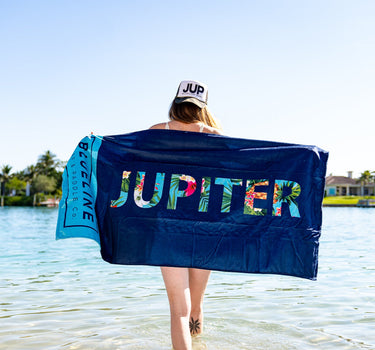 Person holding a navy Blueline Jupiter beach towel at the water’s edge with Jupiter text visible across the towel.