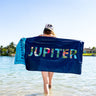 Person holding a navy Blueline Jupiter beach towel at the water’s edge with Jupiter text visible across the towel.